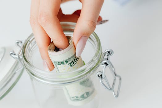 A close-up of a hand placing rolled dollars into a glass jar, symbolizing savings.