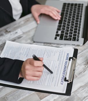 An adult reviews and signs a home insurance policy document on a clipboard next to a laptop.