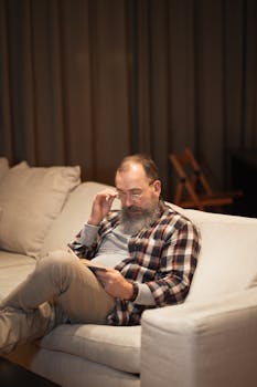 Elderly man sitting on a cozy sofa indoors, using a tablet and wearing glasses.