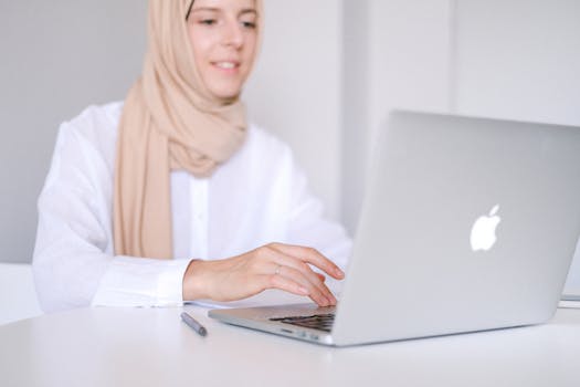 Muslim woman in headscarf smiling while working on laptop at home office.