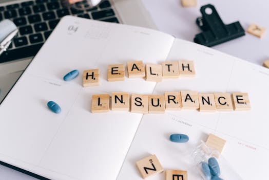 Scrabble tiles spelling 'Health Insurance' on planner with pills and laptop, symbolizing healthcare planning.